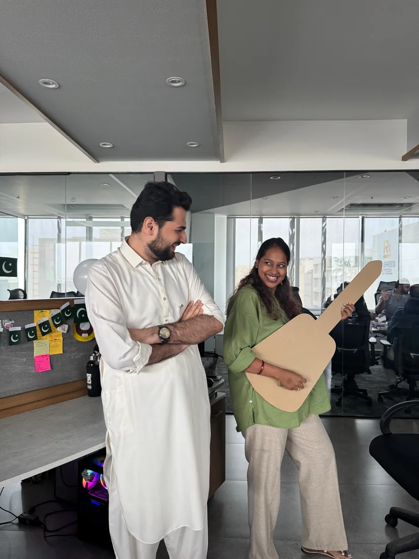 Behind the scenes: two teammates joking around in the office, one holding a cardboard guitar